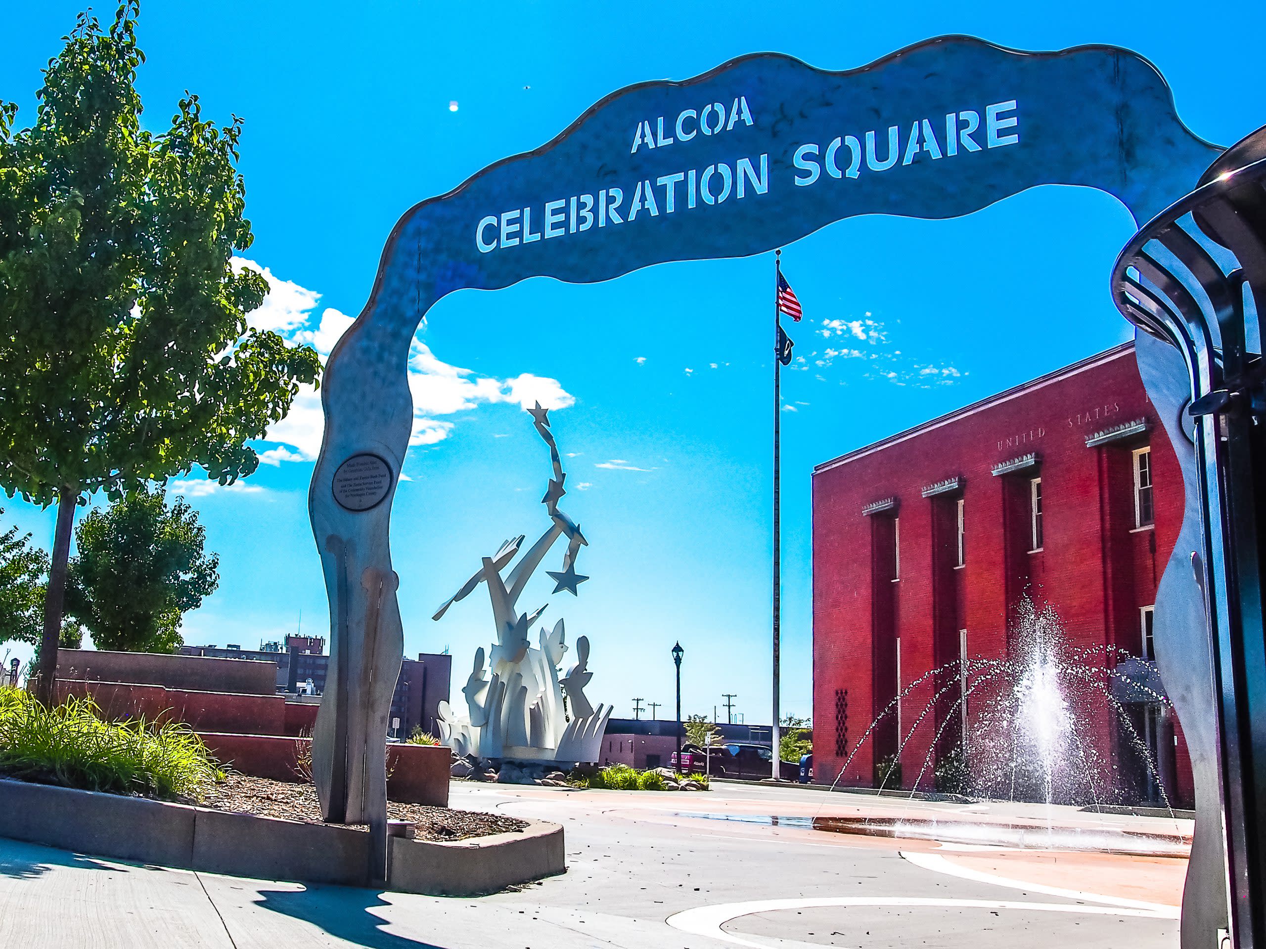 Steel arch entrance with alcoa celebration squre carved at top. red brick building, fountain and white sculpture lay behind. 