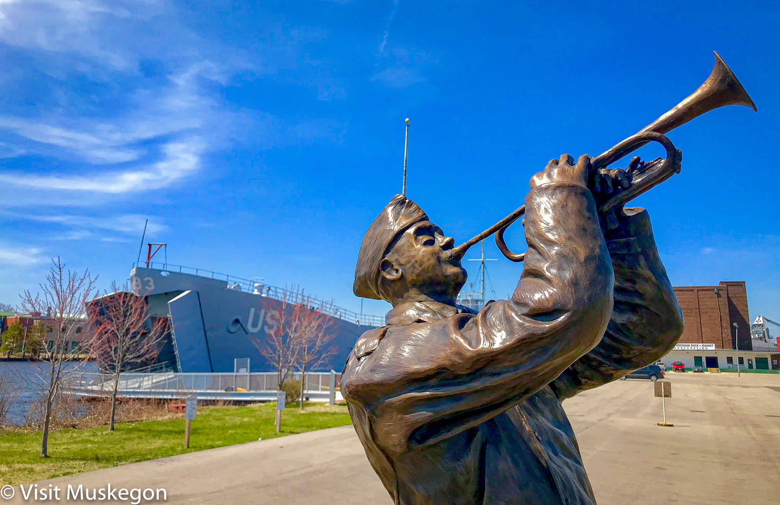 bronze sculpture of world war two soldier blowing trumpet set against blue sky. a world war two landing ship sits in the water behind