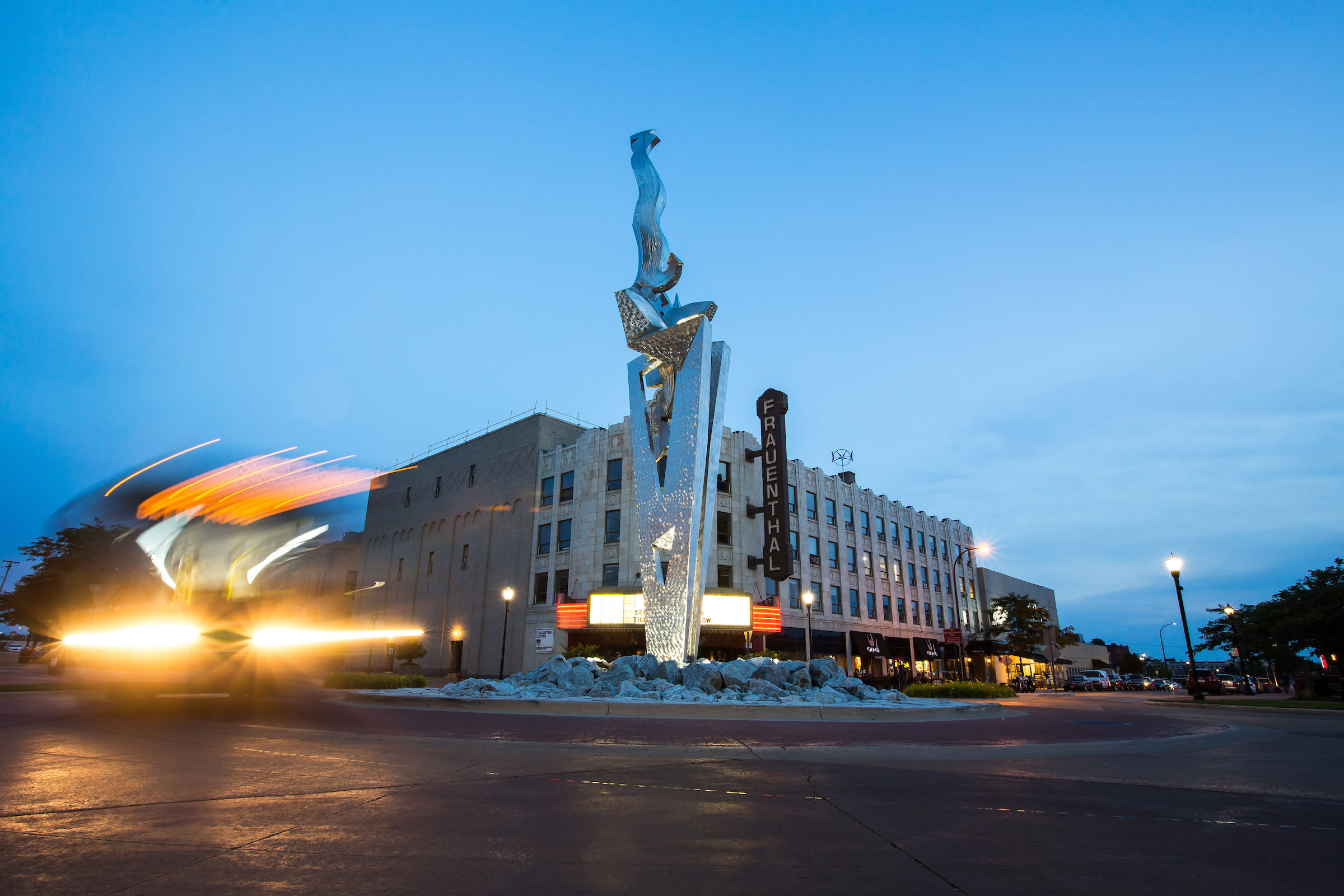 dusky blue sky, large modern steel sculpture pierces intersection in front of historic building with brown sign that reads frauenthal. blurred headlights fro vehicle are to the left of the photo.