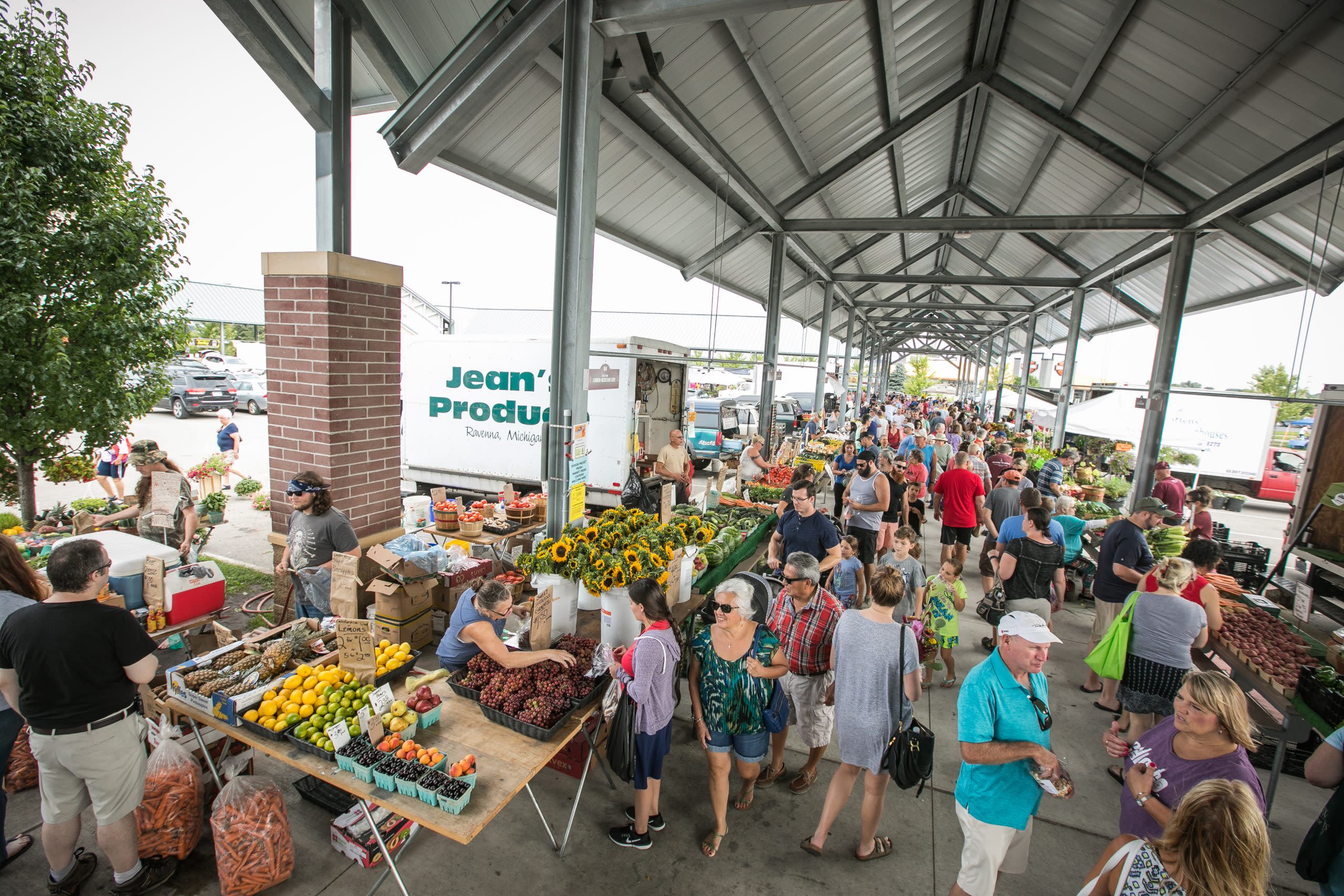 outdoor farmers market shows many shoppers on sidewalk with tables of fresh produce. a vaulted white roof covers them supported by tall white columns