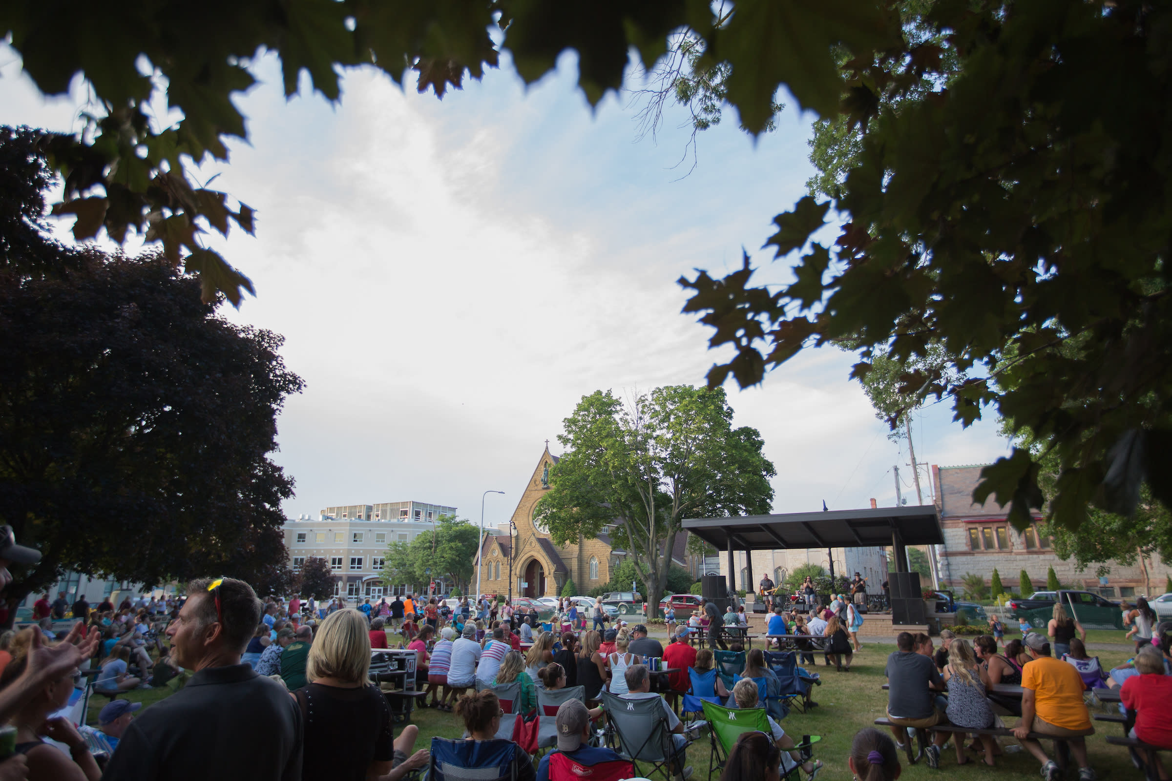 Crowd gathered in outdoor park for summer concert. trees frame photo and outdoor stage is seen in distance