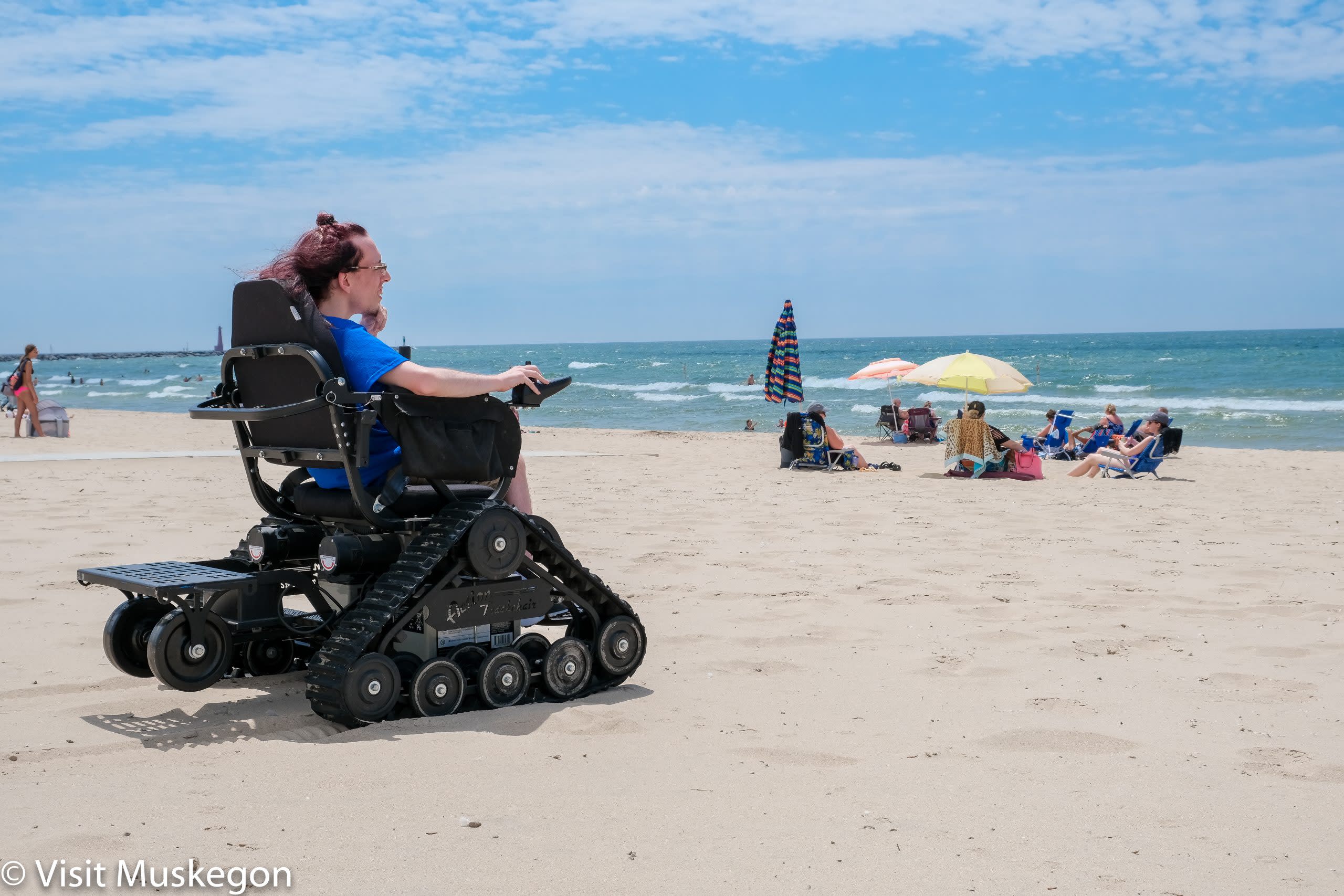 Young man in TrackChair on the beach looks out over Lake Michigan. Beach goers and bright umbrellas are on the shoreline. 