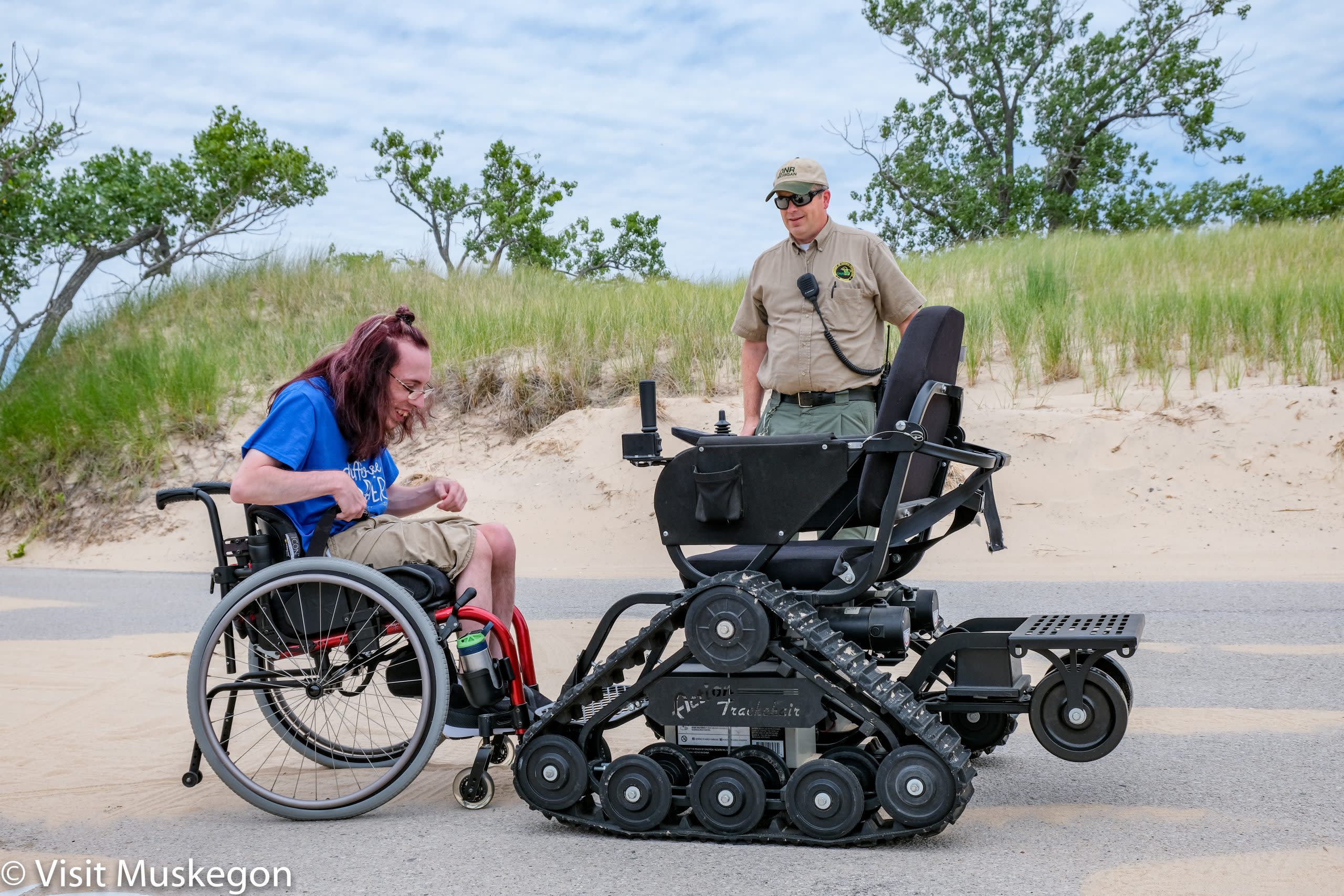 smiling young man in wheel chair sits before a TrackChair. A State park ranger stands nearby. beach dunes and grass are in the background. His blue tee shirt says "difference maker)