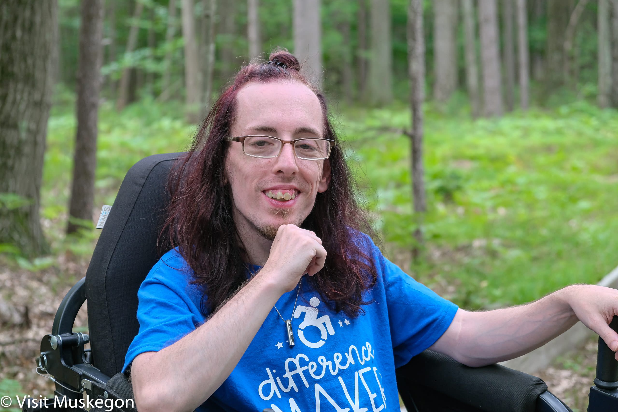 young man holds his fist beneath his chin. he is seated in a TrackChair with ferns and woods in he background. His blue tee shirt says 