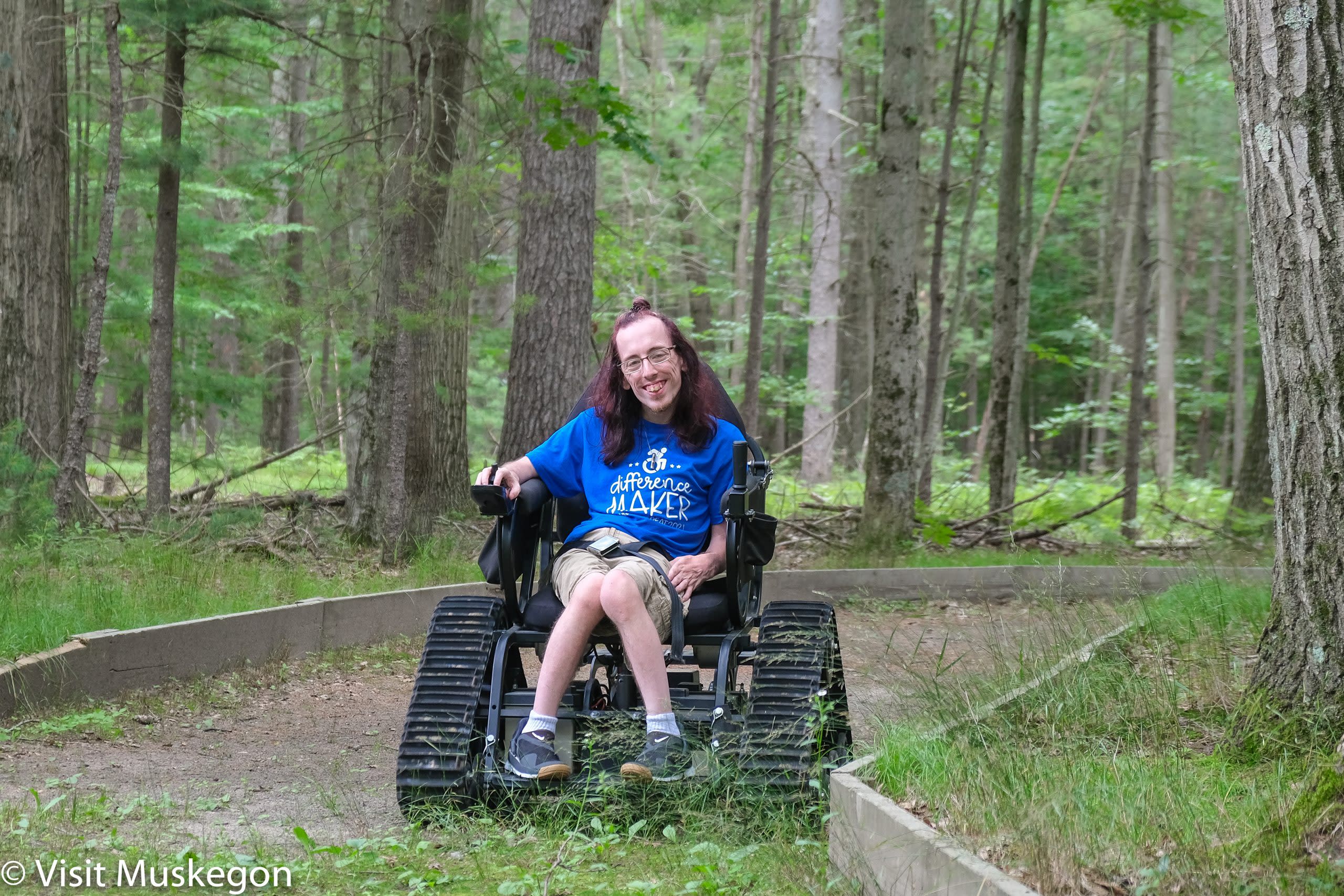 Smiling young man travels hiking path in TrackChair at Muskegon Luge Adventure Sports Park. He is surrounded by trees and natural flora. 