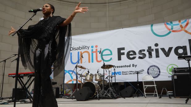 A performer in a sparkling black outfit sings into a microphone on an outdoor stage in front of a large "Disability Unite Festival" banner.