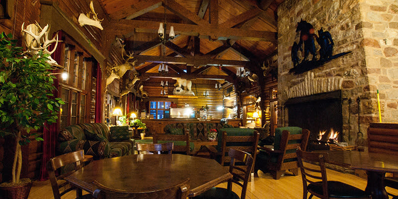 Wooden tables and chairs sit in the main room of a large lodge next to a large stone fireplace