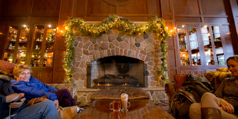 Two women sit in sofas on either side of a stone fireplace adorned with garland at the Osthoff Resort