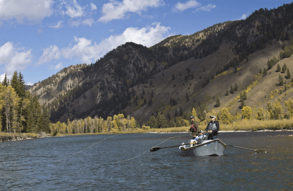 Three travelers cast their lines as two stand and one sits in the boat on an alpine lake in Grand Teton National Park, where the lush mountain range towers in sweeping views behind them. 