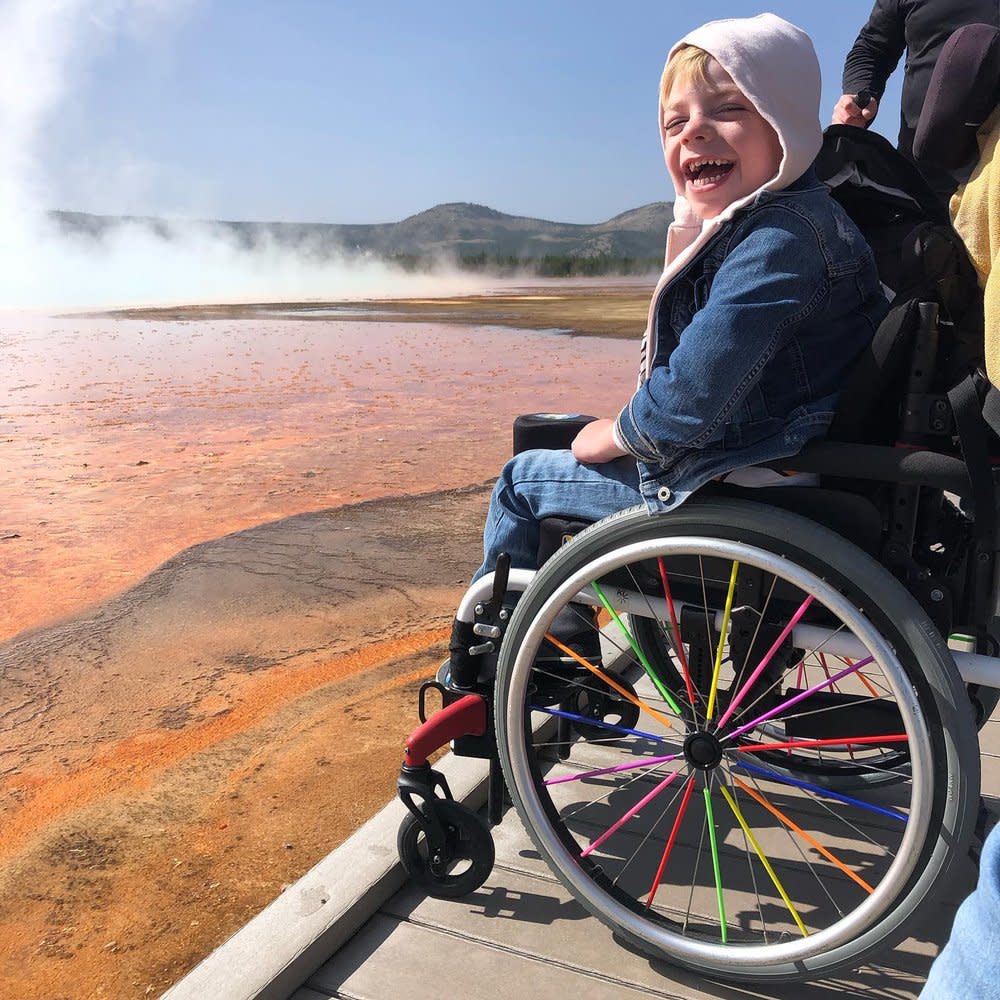 A young child beams a joyful grin in a hoodie and a jean jacket with matching jeans as he sits in a wheelchair looking at the camera, Yellowstone National Park misting behind him. The wheelchair has rainbow-colored spokes.