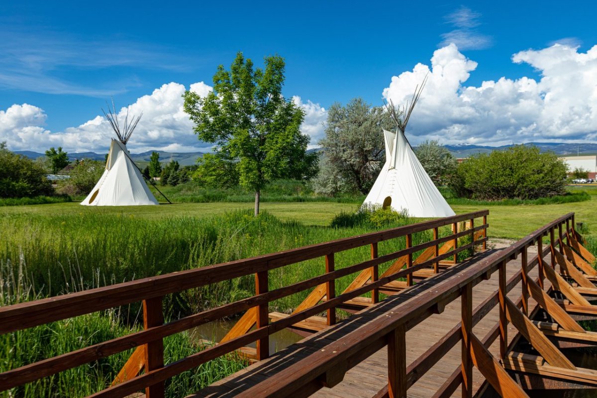 A wooden walkway leads to white teepees standing in Fremont County grasses, with mountains and clouds rising in the distance.