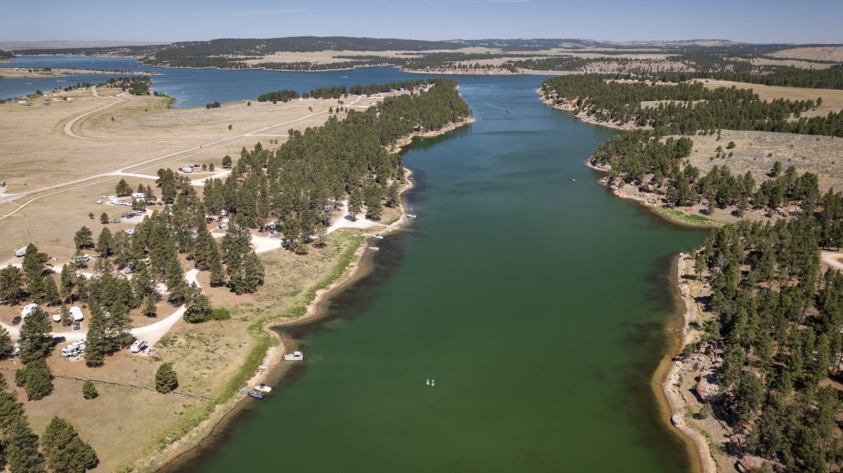 An aerial view shows the winding, teal waters of Keyhole Reservoir framed by pine-covered shorelines and a busy campground dotted with white RVs.