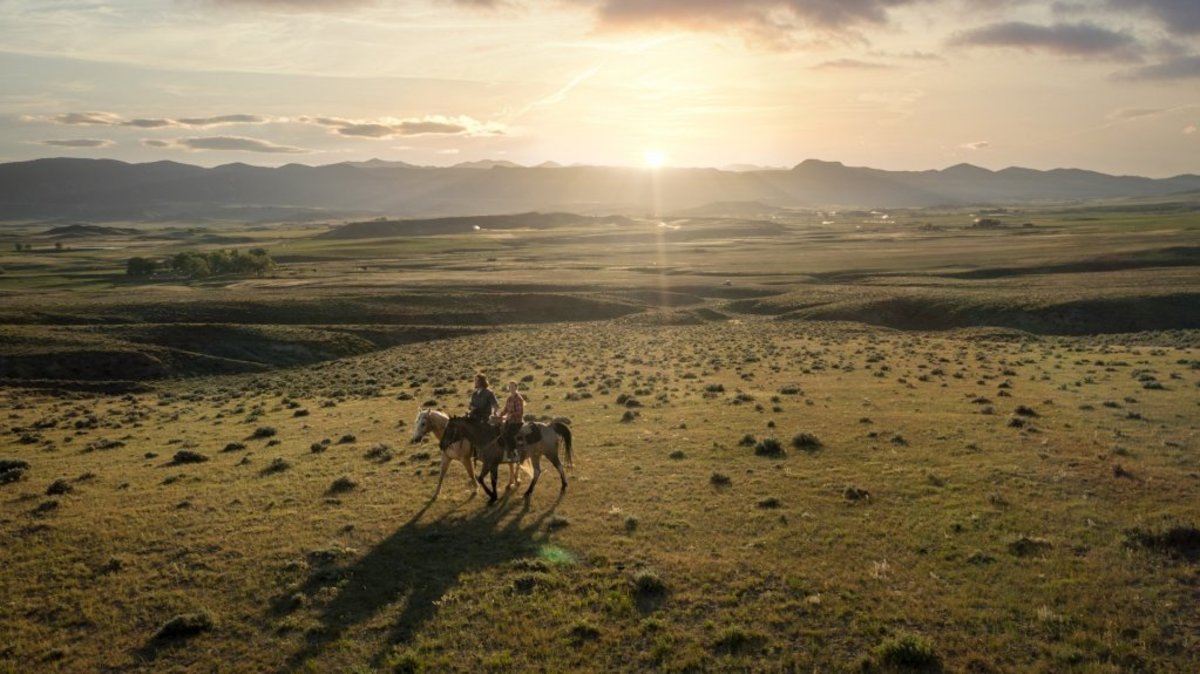 Two people riding horses across a sprawling open landscape with mountains in the background at TA Guest Ranch.
