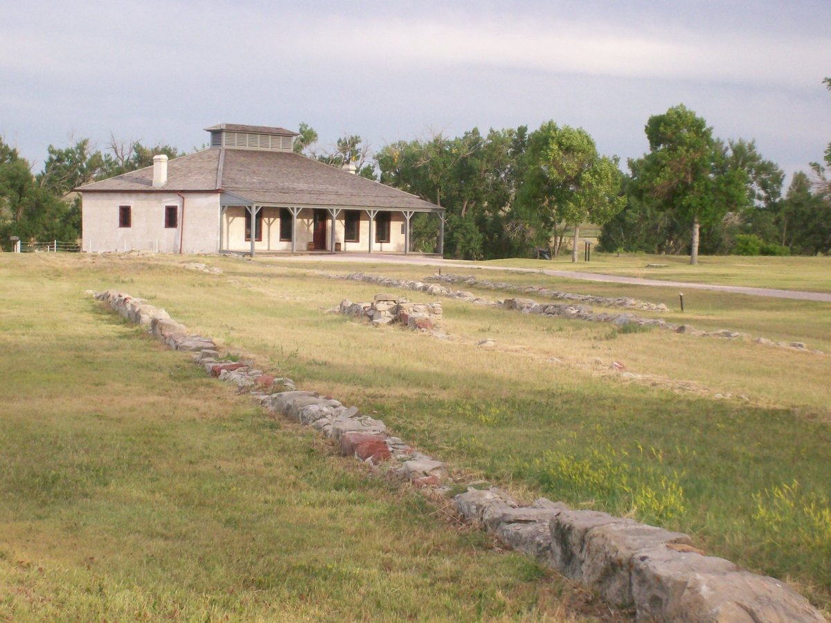 Historic stone foundations leading to a preserved 19th-century military building surrounded by trees and open prairie.