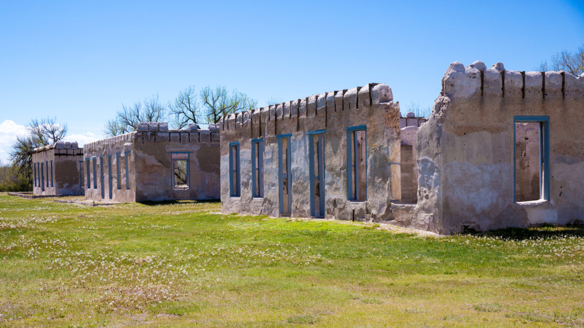Crumbling adobe and stone ruins of historic Fort Laramie under a clear blue sky with scattered trees and green grass.