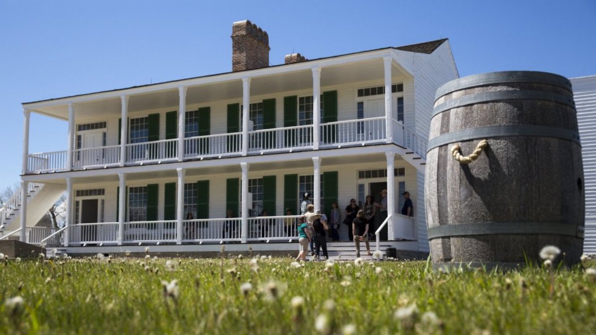 A wooden barrel in the foreground and a group of people on the steps of the Old Betlam building in the background at Fort Laramie.