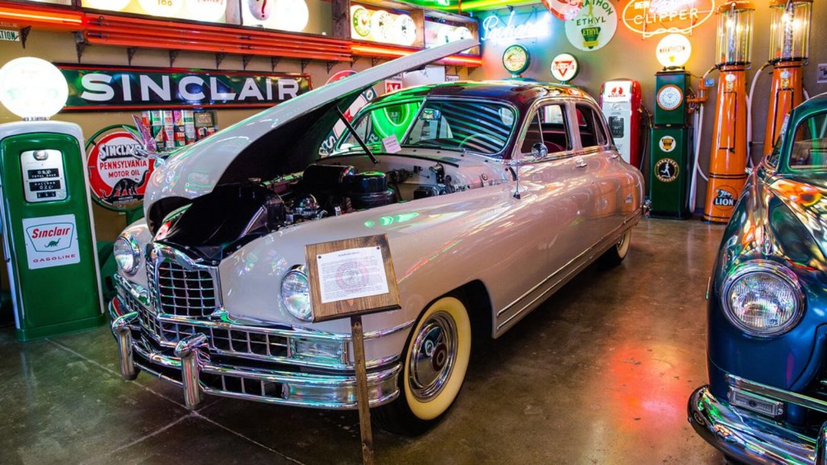 An old-fashioned car with the hood raised surrounded by old-fashioned gas pumps and neon signs at the Frontier Auto Museum.