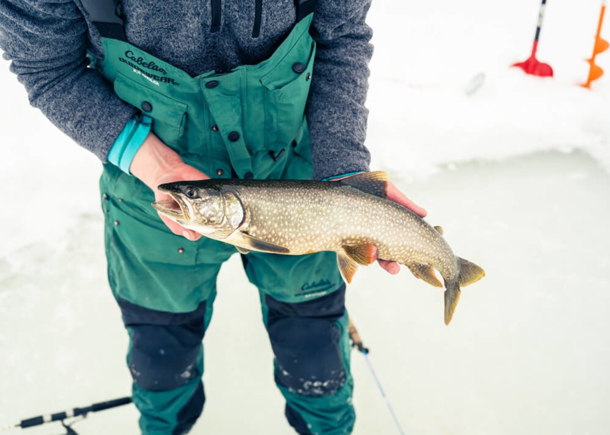 An ice fisherman in winter gear shows off a fish caught in a nearby hole.