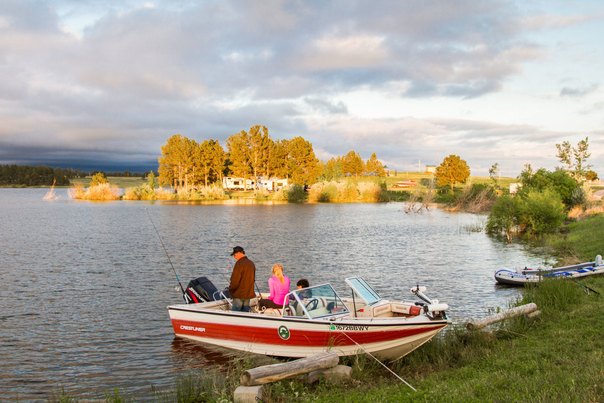 A man stands as his daughter sits next to him, with his son to the right of his daughter, on a Crestliner fishing boat, poles in hand, overlooking the waters and wilderness in Wyoming.