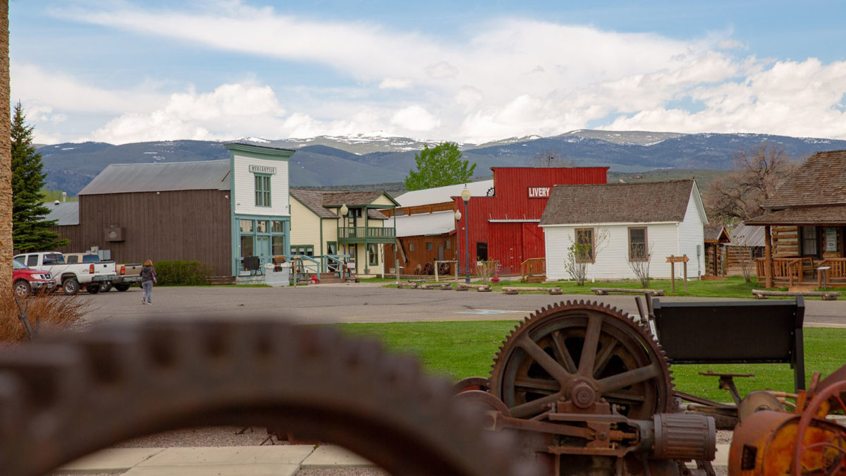 Overlooking a ranch in Wyoming, where a woman walks atop the pavement, heading to the ranch buildings as snowcapped mountains rise in distance beneath the clouds.