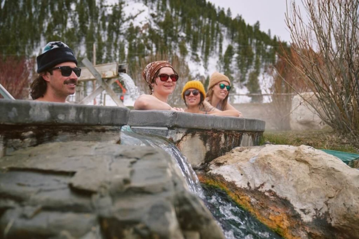 A family wearing hats (one in a bandana) relax at the Astoria Hot Springs and Park, in sunglasses and all smiles before a snowy landscape behind them.
