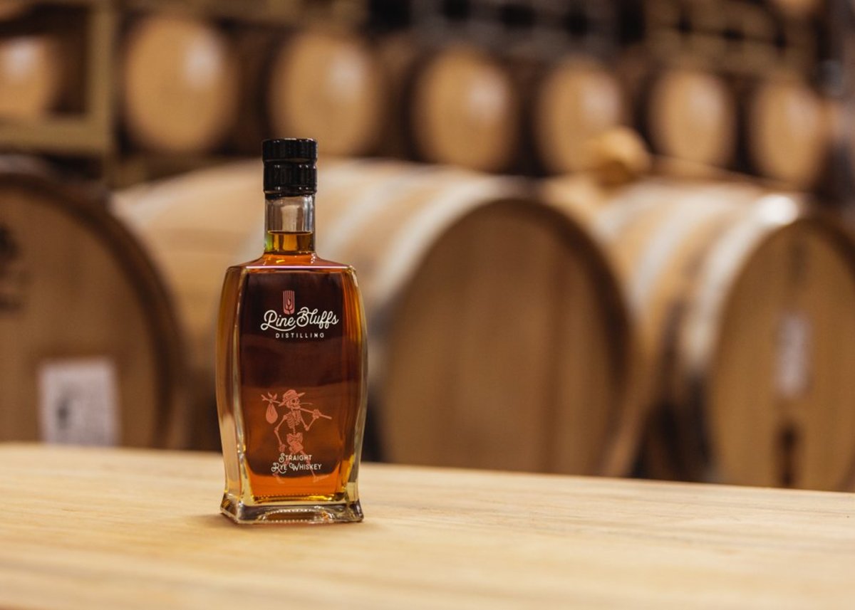 An amber bottle of Straight Rye Whiskey, a craft whiskey distilled at Pine Bluffs Distilling in Pine Bluffs, Wyoming, stands atop a wooden bar, with oaks barrels lining the background.