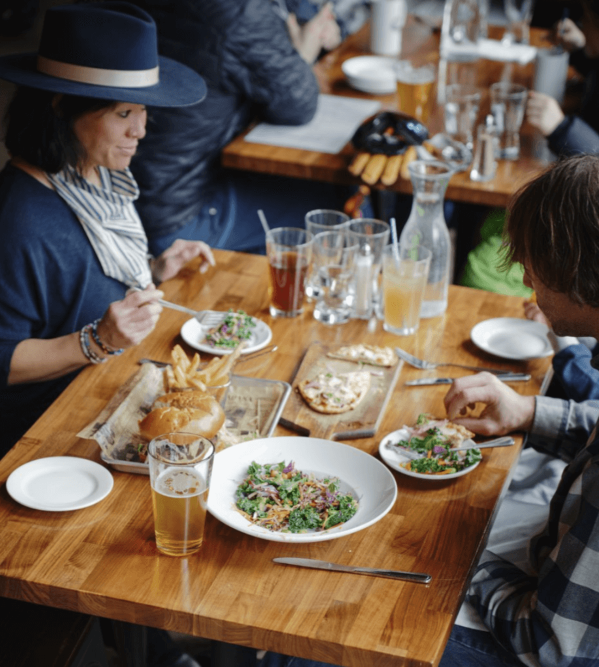 Two people seated at a table with several glasses, plates of salad, a wood-fired pizza and a newspaper lined tray holding a burger and fries inside of a restaurant.