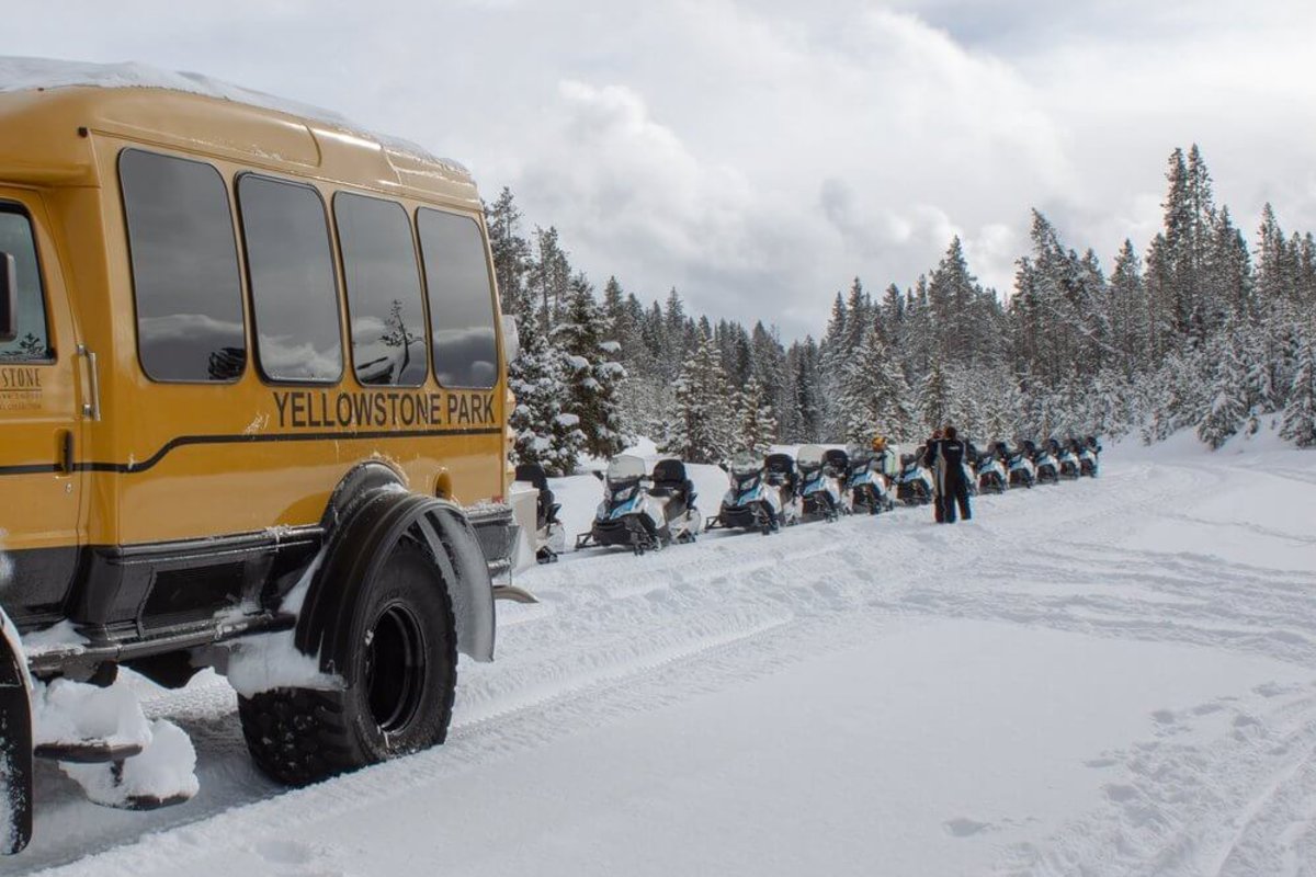A yellow bus with snowmobiles behind it amongst a snowy white landscape in Yellowstone National Park during the winter.