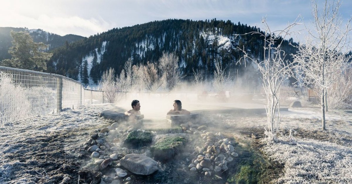 A couple soaking in a pool at Astoria Hot Springs, surrounded by a snow-covered landscape and a snow and tree-covered mountain in the distance.