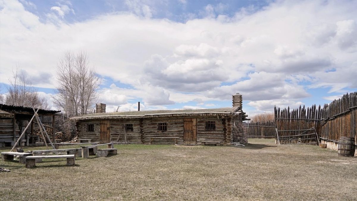 Historic homes sit in a grassy field, with a flag waving in the distance above the mountain ridge.