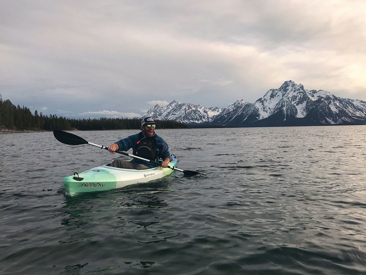 A man goes kayaking by Jackson Lake Swim Beach, as the Teton Range towers in the background.