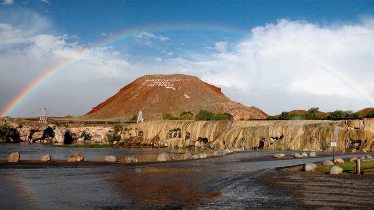 A rainbow appearing over a large rock formation with a sign that reads, "World's Largest Mineral Hot Spring" in the background and a road leading past a series of smaller rock formations with water flowing over them in the foreground at Hot Springs State Park.