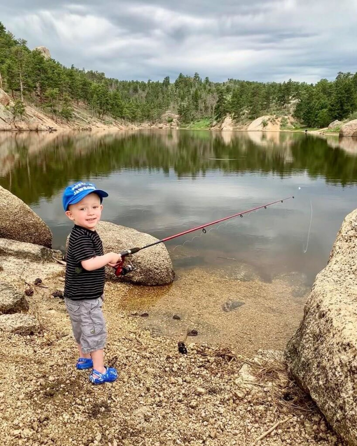 A happy young boy wearing a baseball cap holds a fishing rod, ready to cast into the waters of Curt Gowdy State Park in Laramie County, Wyoming, as trees reflect in the water.</p>
<p>