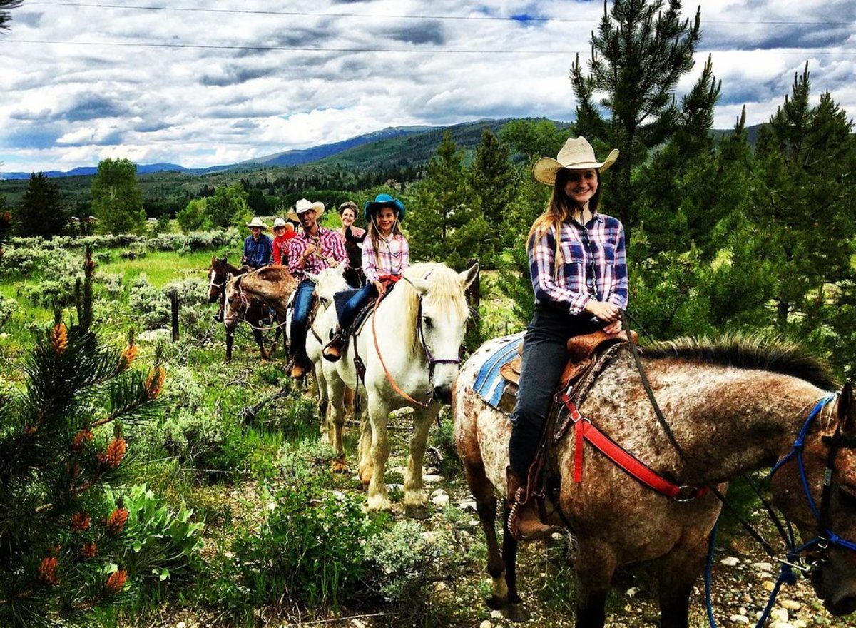 A group of people riding horses in a single file line on a trail surrounded by trees and shrubs at Medicine Bow Lodge and Adventure Guest Ranxh.