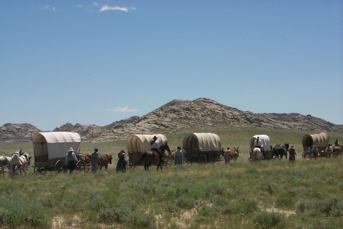 People stand beside covered wagons and horses in tall grass, with rugged mountains rising in the distance.