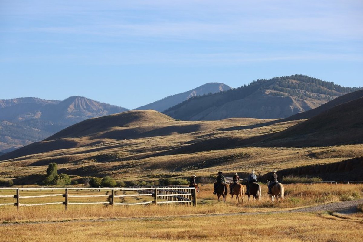 A group of people riding horses past a wooden fence through an open field surrounded by hills and mountains at Red Rock Ranch.