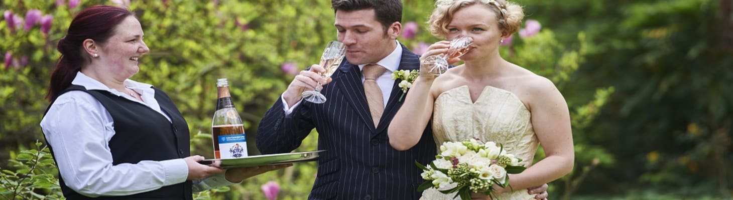 A bride and groom being served a glass of champagne in the garden at Lucy Cavendish College.