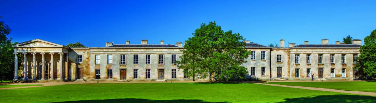 A landscape external view of the Georgian style building showing a clear blue sky, green lawn and tree in the center.