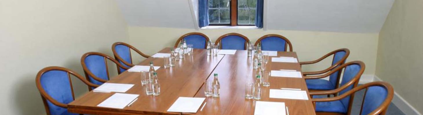 Small syndicate room with natural daylight and pale walls. Large wooden boardroom table with blue chairs, perfect for smaller meetings, interviews or as a break-out area.