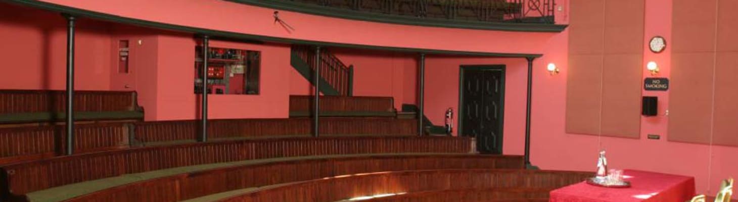 Dark salmon pink decor Victorian lecture room, with continuous upholstered bench seating in two tiers across a curving auditorium, a raised stage and gallery. Perfect cambridge university venue hire for lectures, conferences and events.