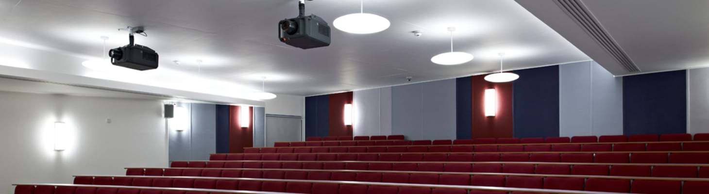 A contemporary designed large lecture theatre with white walls, rows of red uniform chairs and two over head projectors.