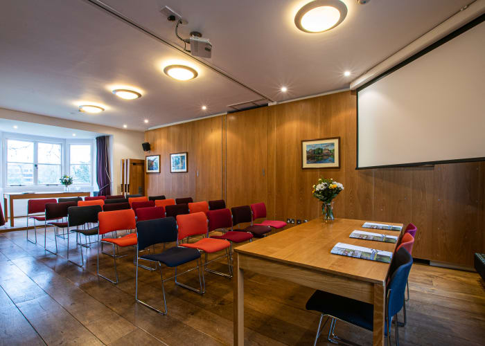 A wooden panelled meeting room filled with natural daylight with a large screen and chairs set theatre style.