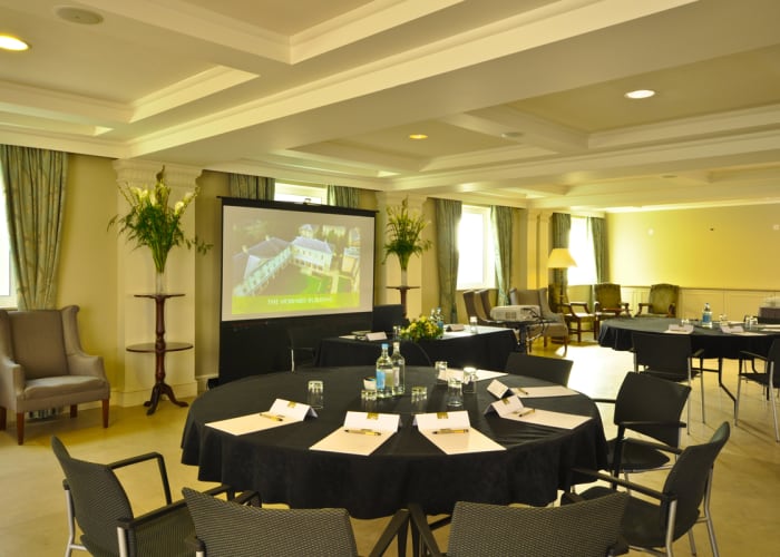 Large and light meeting room. Tables set up cabaret style, black table cloths and chairs. Projector screen in central view.