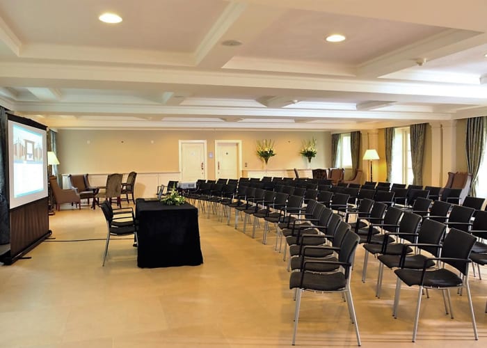 A large cream room set up with rows of black chairs in front of a speaker desk and projector screen, ideal for day meetings.