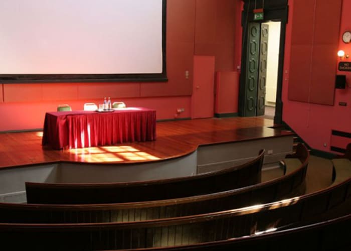 Dark salmon pink decor Victorian lecture room, with continuous upholstered bench seating in two tiers across a curving auditorium, a raised stage with top table. Perfect Cambridge university venue hire for lectures, conferences and events.
