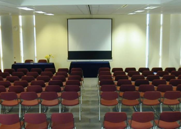 Bright room with a large screen on the wall, room has a top table for two and numerous dark red rows of chair set theatre style
