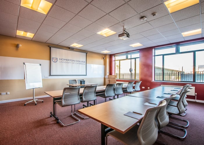 Bright spacious room with two large windows on one side of the room, set up with two long tables with chairs and fully equipped with AV equipment, including a screen and flipchart