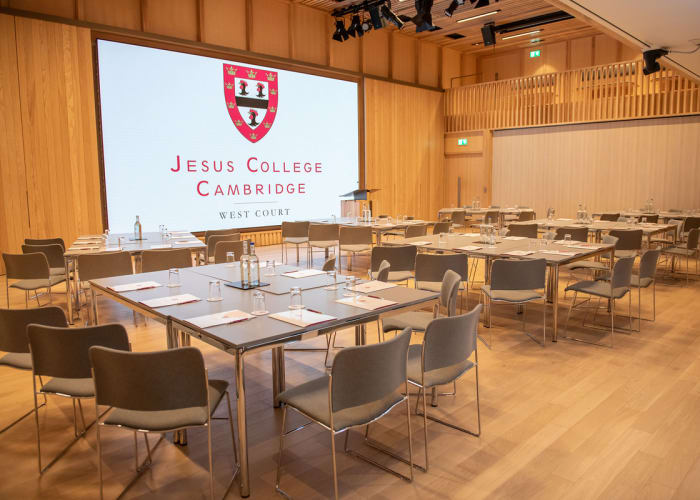 Full wood hall with cabaret style seating in front of a large screen showing Jesus College logo at a closer view