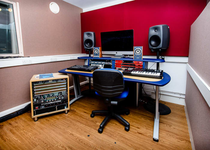 A small fully equipped room for music making with red and pink walls, speakers, computer, blue desk and chair for room hire