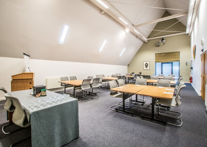 Modern meeting room with a slanted angled ceiling, bright space with four square tables with chairs around each table, there is also a top table and lectern.
